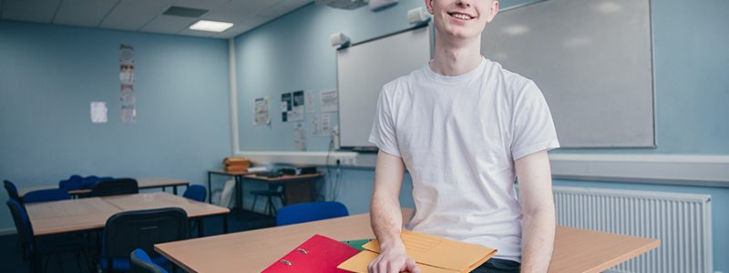 A redhead young man in casual clothing looks at the camera as he stands in an engineering classroom.